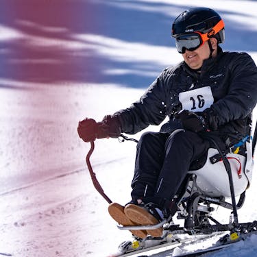 A man smiles while riding a bi-ski, with a guide trailing behind