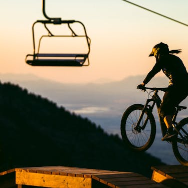 Bike rider getting air over a wooden obstacle with a chairlift and sunset in the background