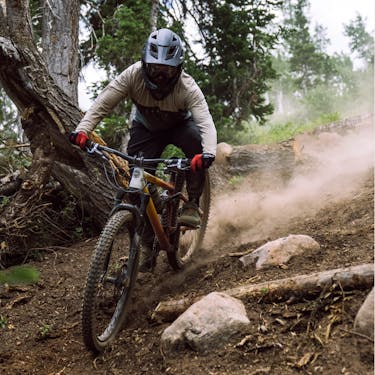 A mountain biker rides past logs and rocks leaving behind a dust trail