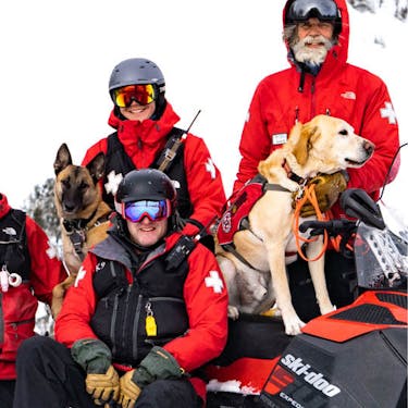 A group of ski patrol team members with ski patrol dogs sitting on snowmobiles