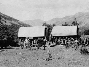 Two parked covered wagons on dirt with mountains in the background