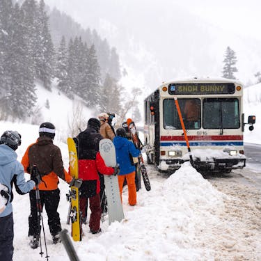 A white Powder Mountain shuttle parked with a line of skiers and snowboarders waiting in line to board
