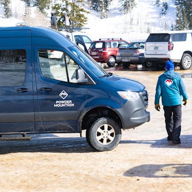 A navy Powder Mountain sprinter van parked in the lot with a Powder Mountain employee walking in front of it