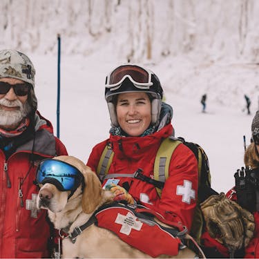 Three ski patrol members smile accompanied by a yellow lab ski patrol dog wearing goggles