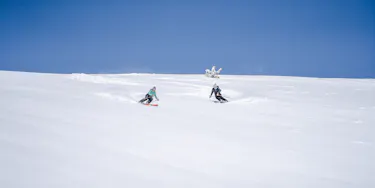 Two skiers skiing down an untouched field of snow with no trees. The sky is bright blue with no clouds.