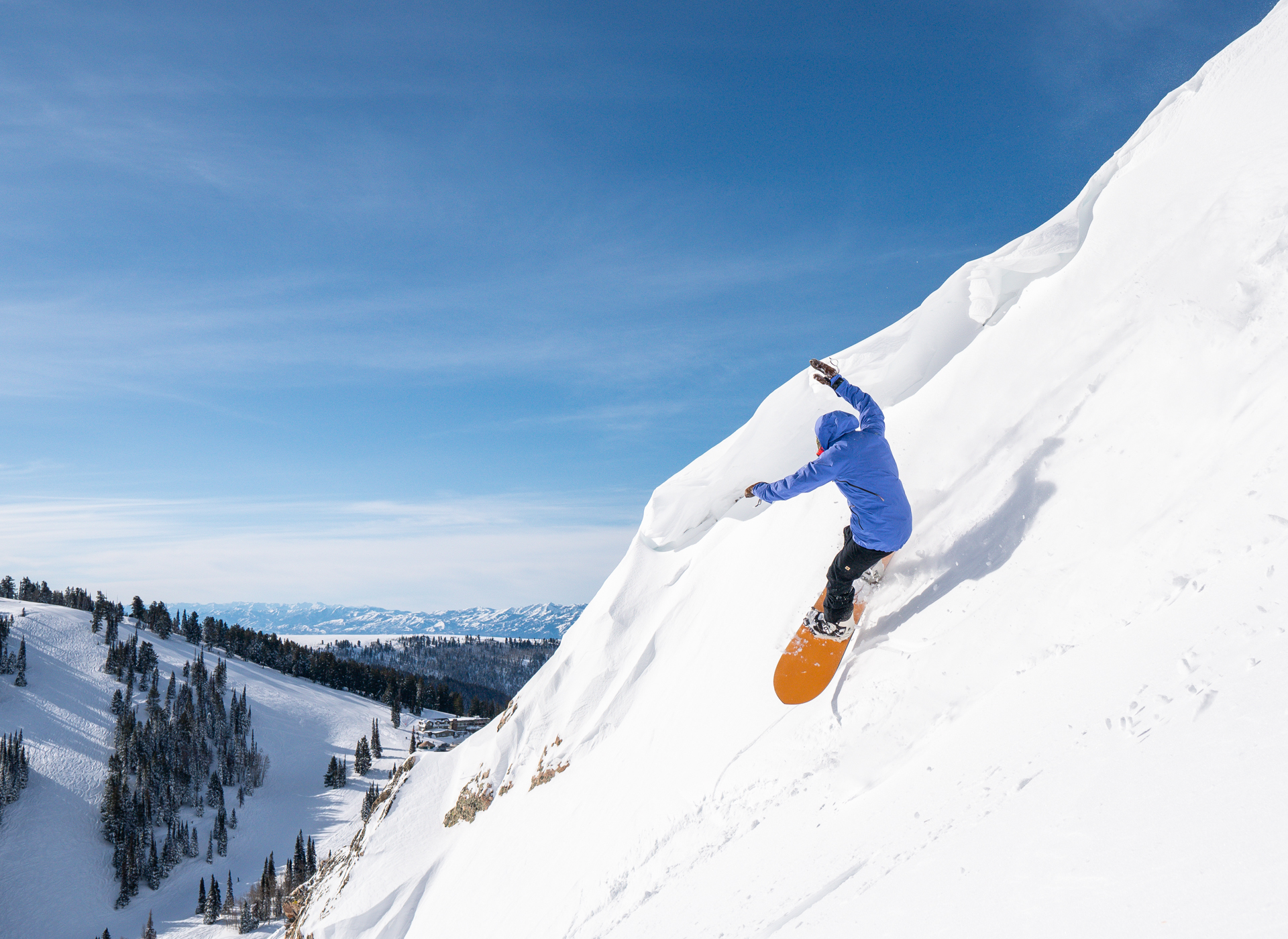 Snowboarder turning on a very steep pitch of snow. They have a bright blue jacket on and an orange board.