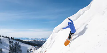 Snowboarder turning on a very steep pitch of snow. They have a bright blue jacket on and an orange board.