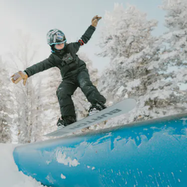 Young snowboarder jumping onto a blue metal tube in the park. The trees in the background are covered in snow.