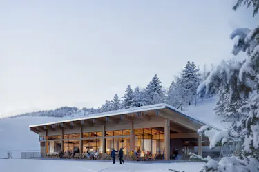 Concept of the new Sundown Lodge at Powder Mountain. The building is large and modern with big windows and natural wood walls. It has a patio in front of the building. The building, ground and trees are snow covered.