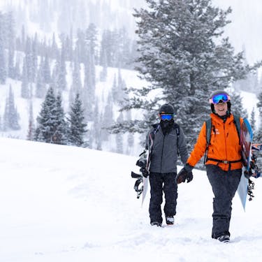 A smiling Powder Mountain guide leads a snowboarder across the snow with boards in hand