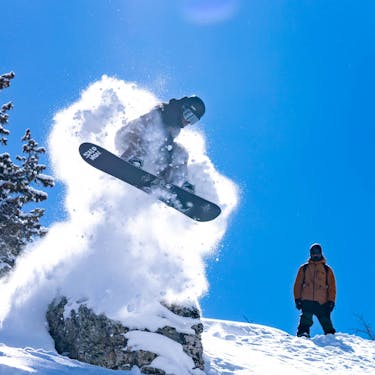 A snowboarder launches off of a boulder with a cloud of trailing snow as another snowboarder watches on