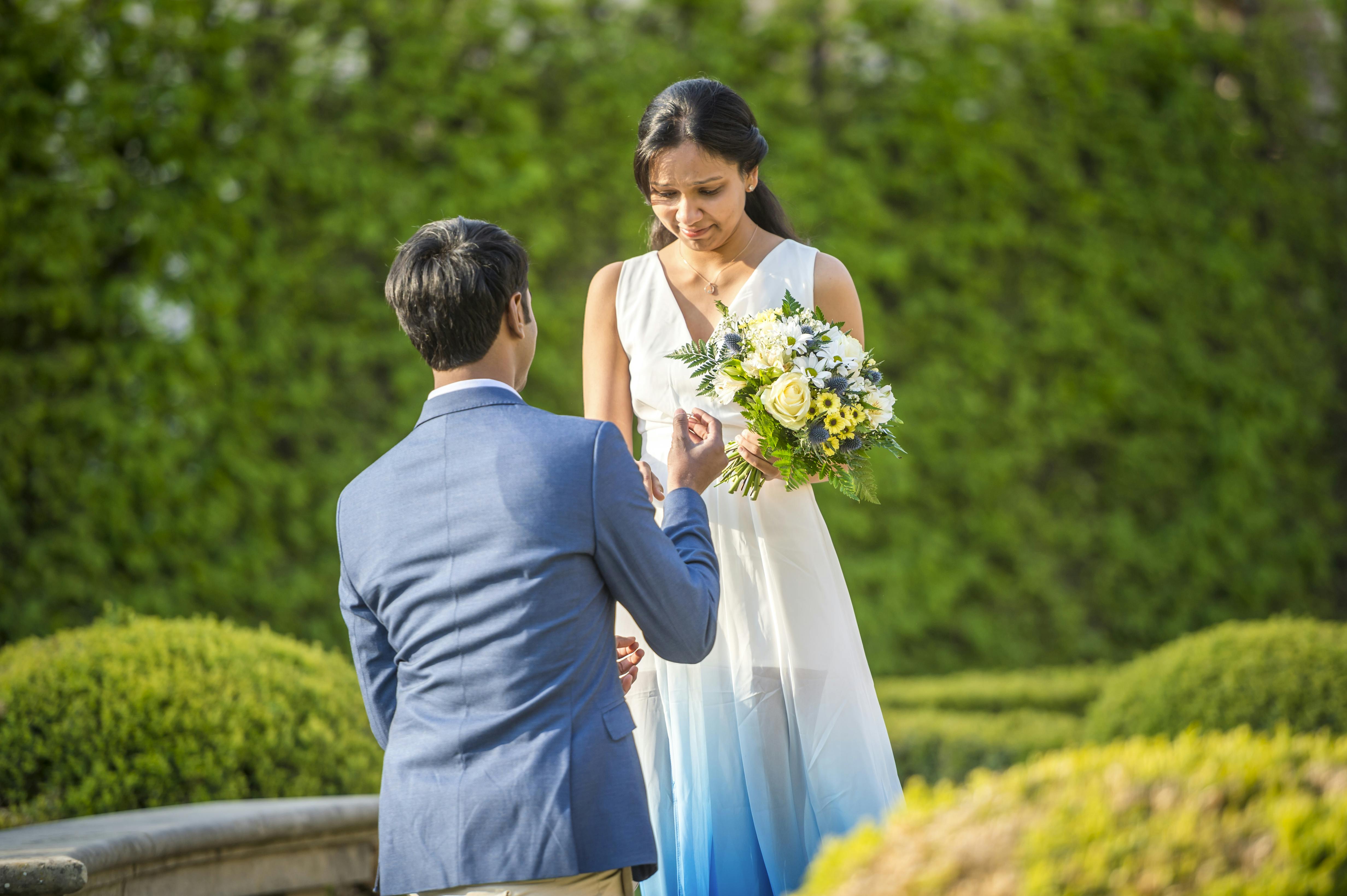 A man on one knee presenting a ring to his fiancée holding a bouquet, surrounded by lush green hedges in a Baroque garden.