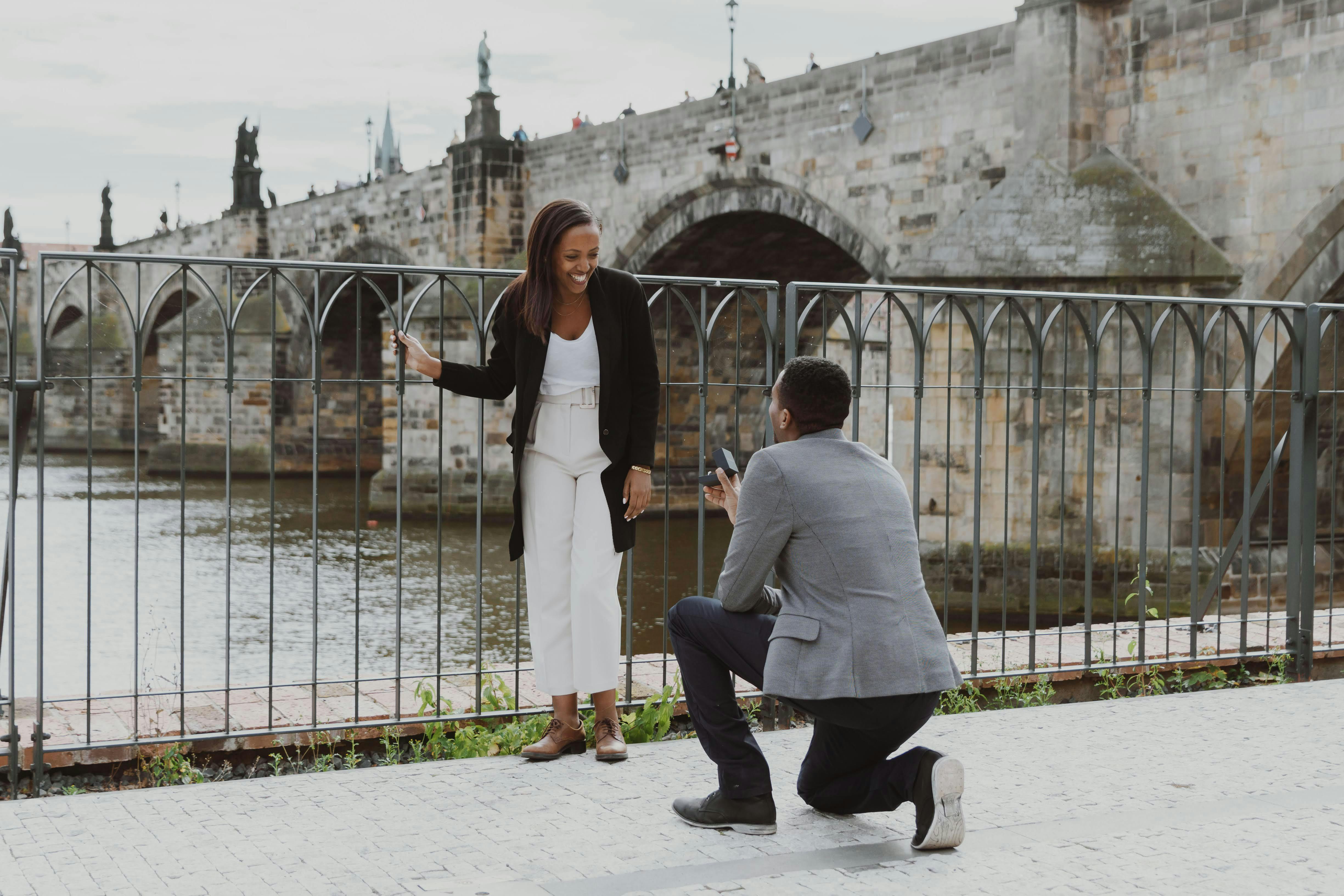 A man kneels to propose to his partner by the Vltava River with Charles Bridge in the background at dusk.