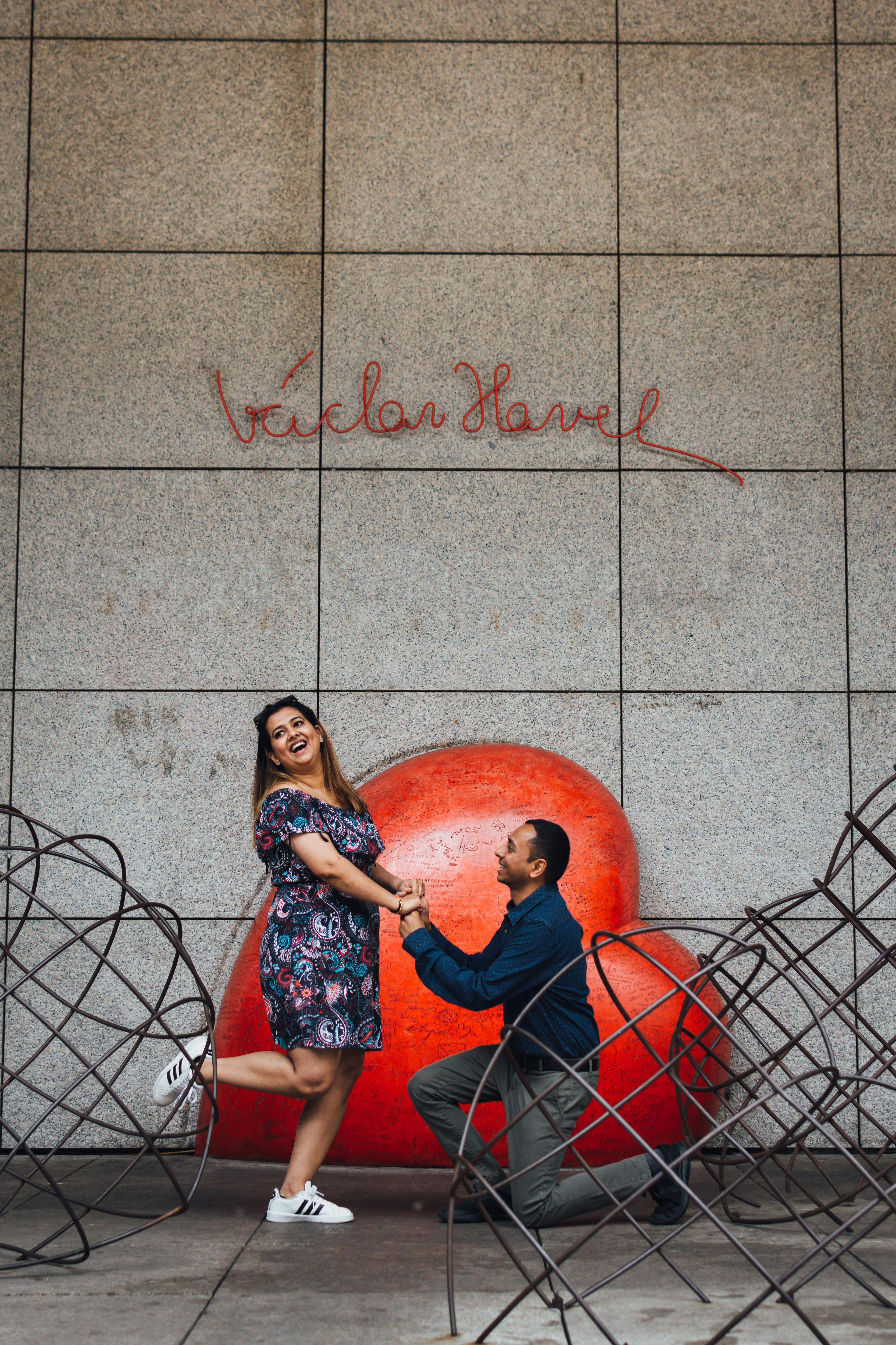A man kneels to propose in front of a large red heart sculpture on a city street, with artful wire benches surrounding the couple.
