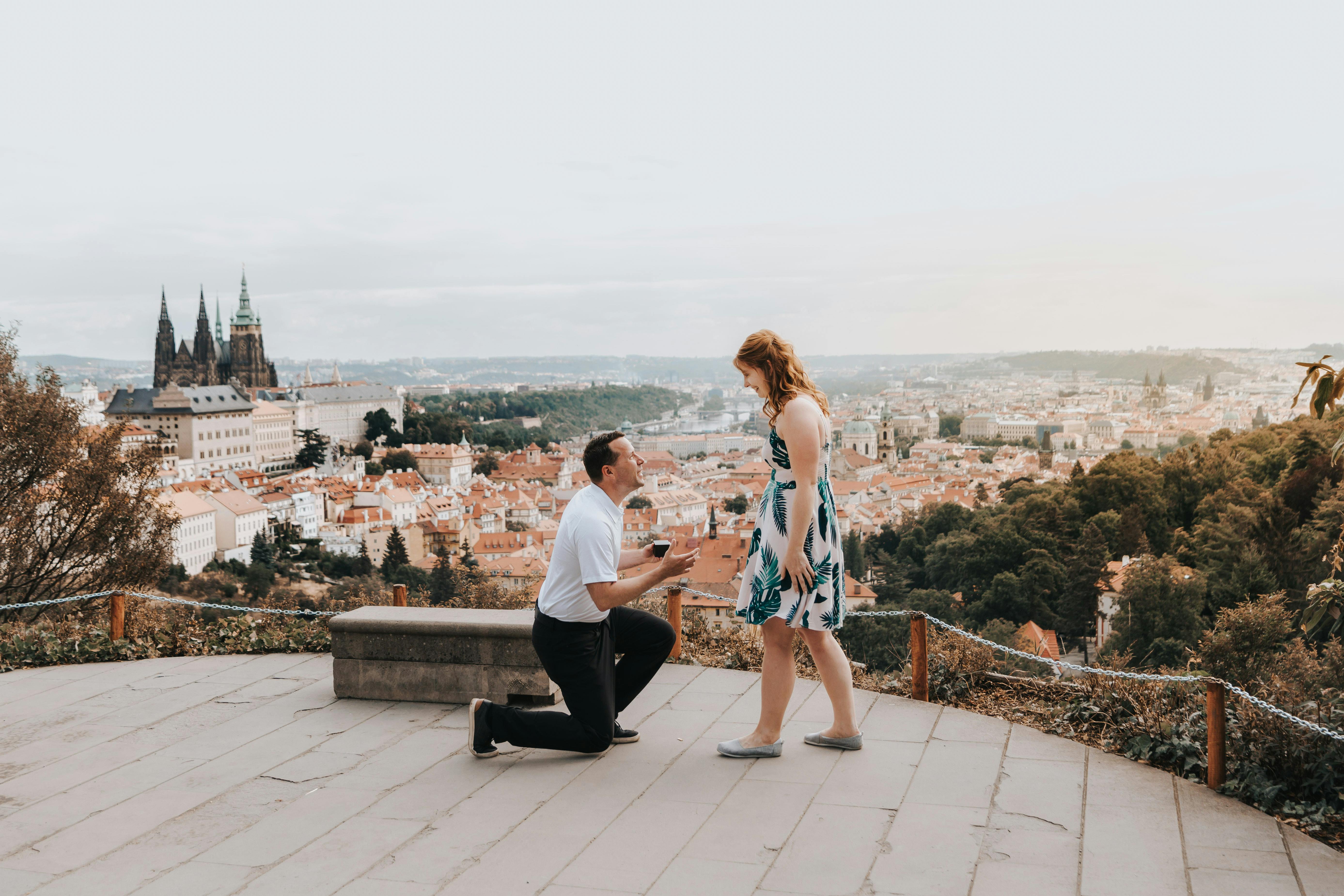 A man on one knee proposing to his partner on a hill terrace with Prague’s red rooftops and castle in the background at sunset.
