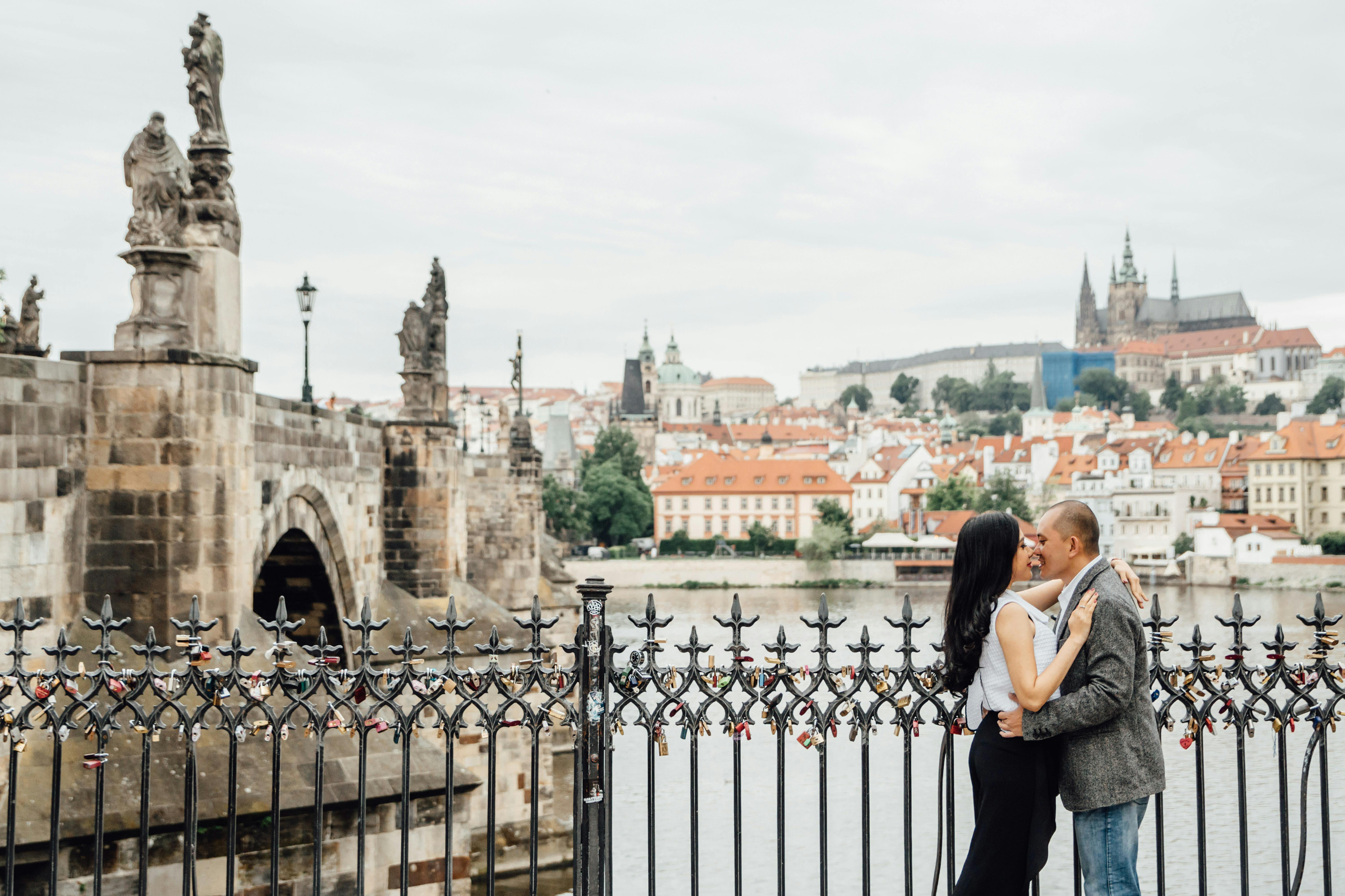 A couple hugs by an ornate railing on the Vltava Riverbank with Charles Bridge and Prague Castle in the background.