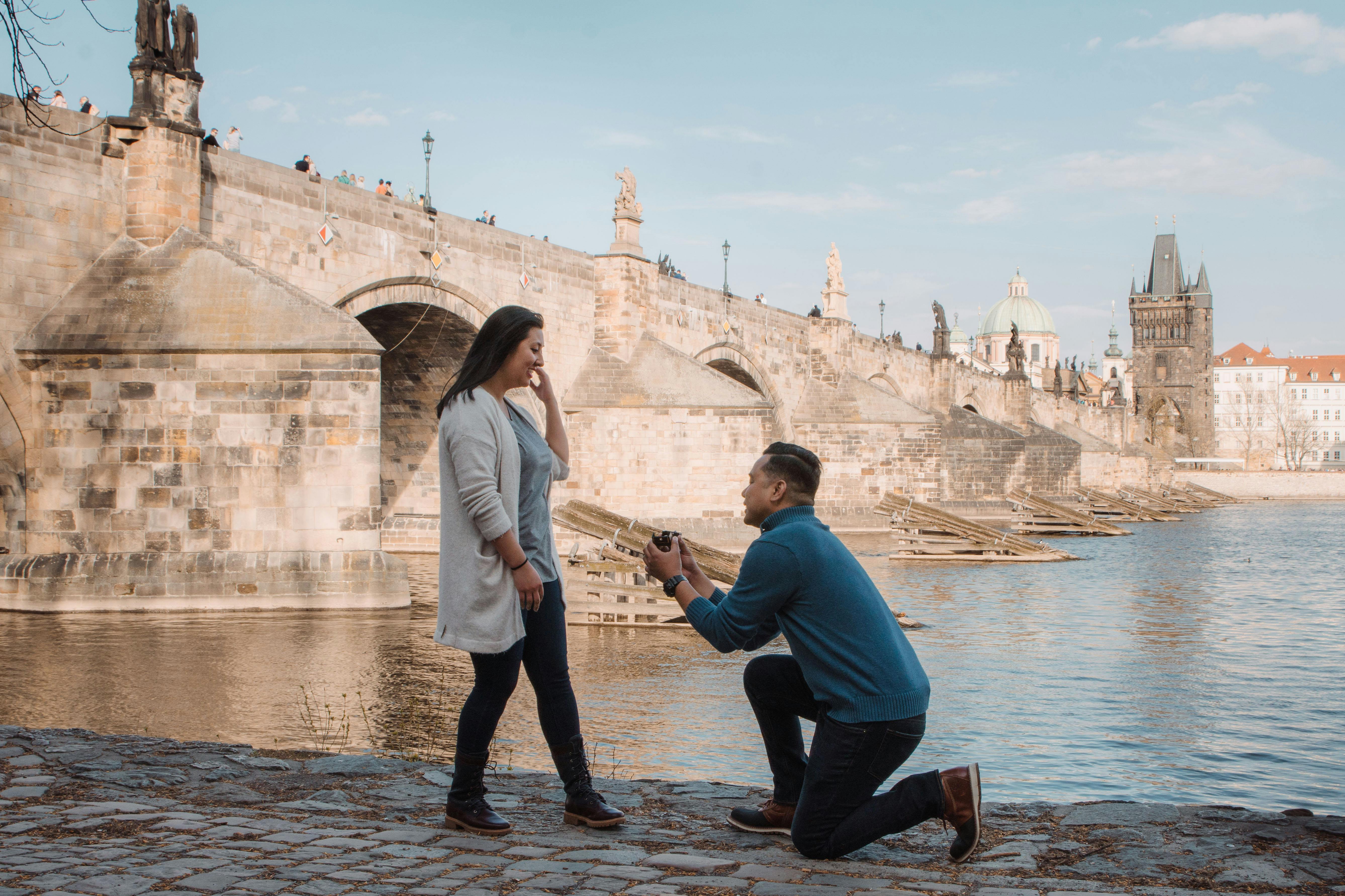 A man kneels to propose to his partner on the riverbank with Charles Bridge and Prague Castle in the background under a clear sky.