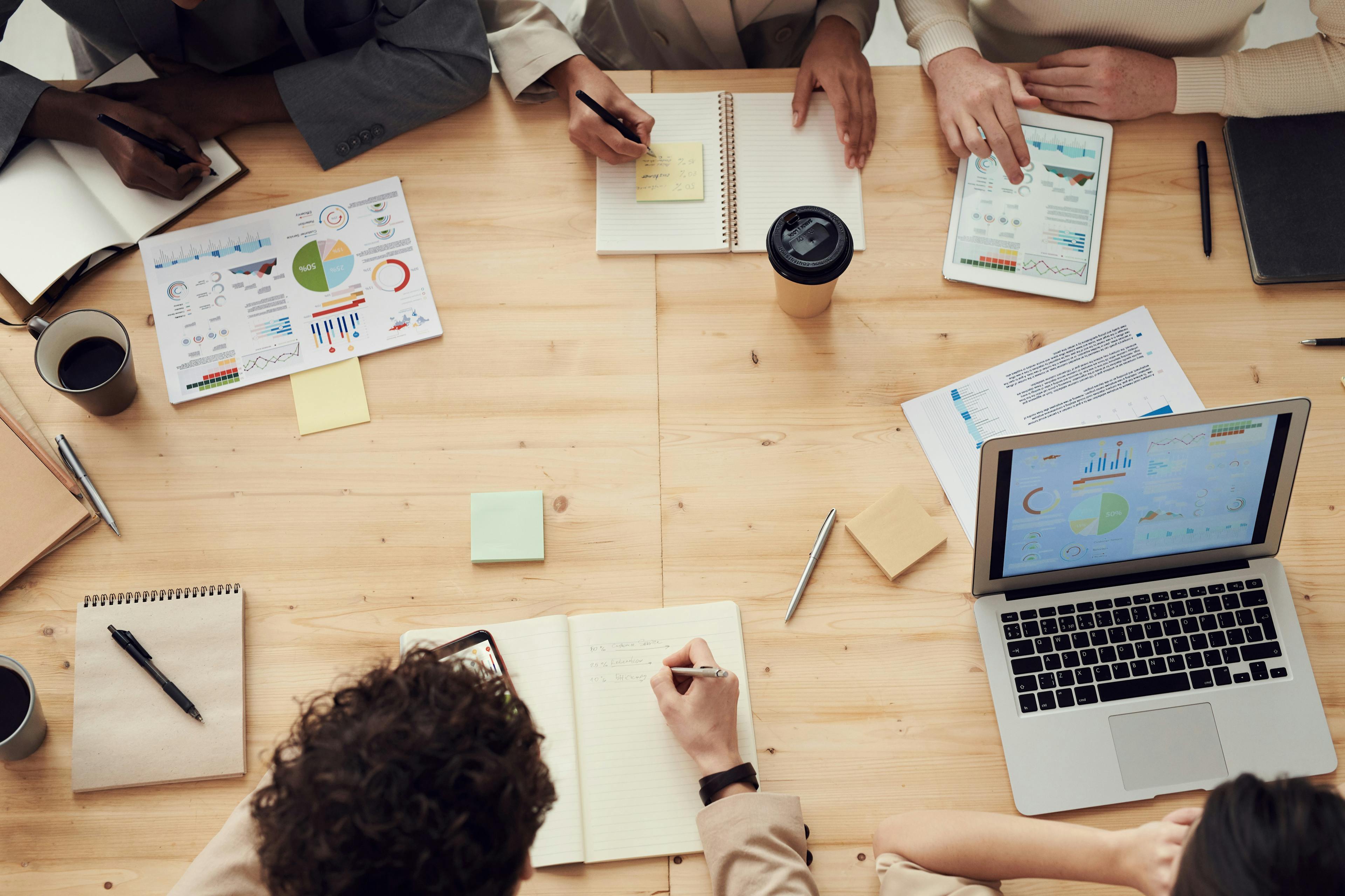 An image of a team meeting around a table that is scattered with company reports, notebooks, a laptop, and an iPad.