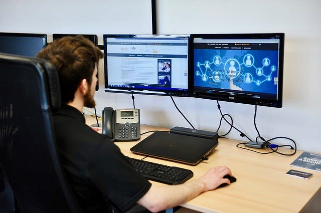 Person sitting at a light wood desk with computer monitors and a laptop.