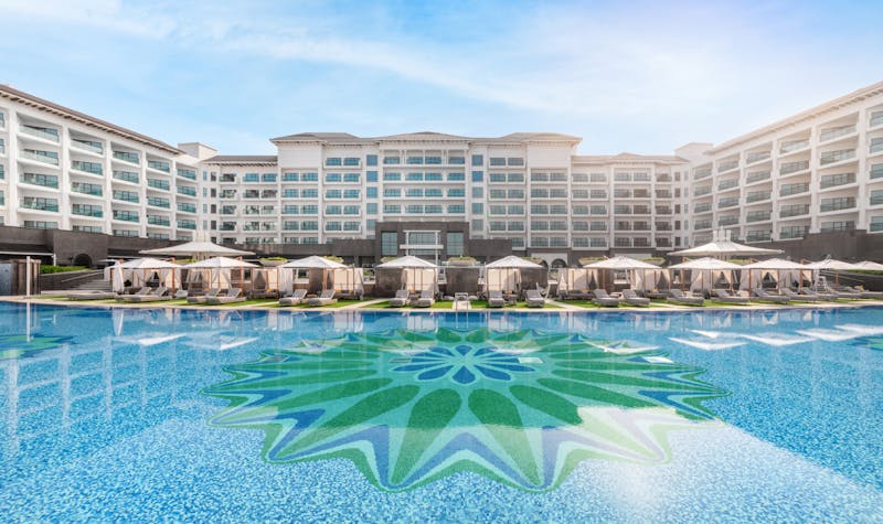 Hotel building behind a large swimming pool reflecting the sunlight, surrounded by shaded cabanas. 