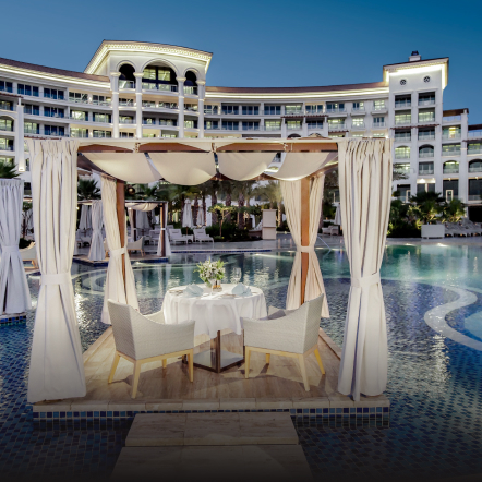 Shaded poolside cabana with tables and chairs, in front of a hotel building. 