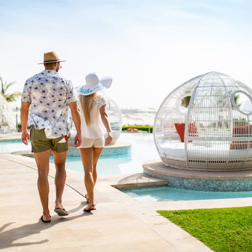 Male and female walking by the pool, with crystal-clear water reflecting sunlight and surrounded by cabanas at the W Dubai The Palm, UAE.