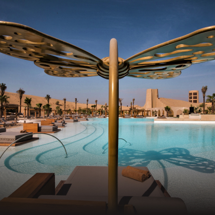 Sun lounger overlooking a swimming pool with crystal-clear water reflecting sunlight, beneath a blue sky. 