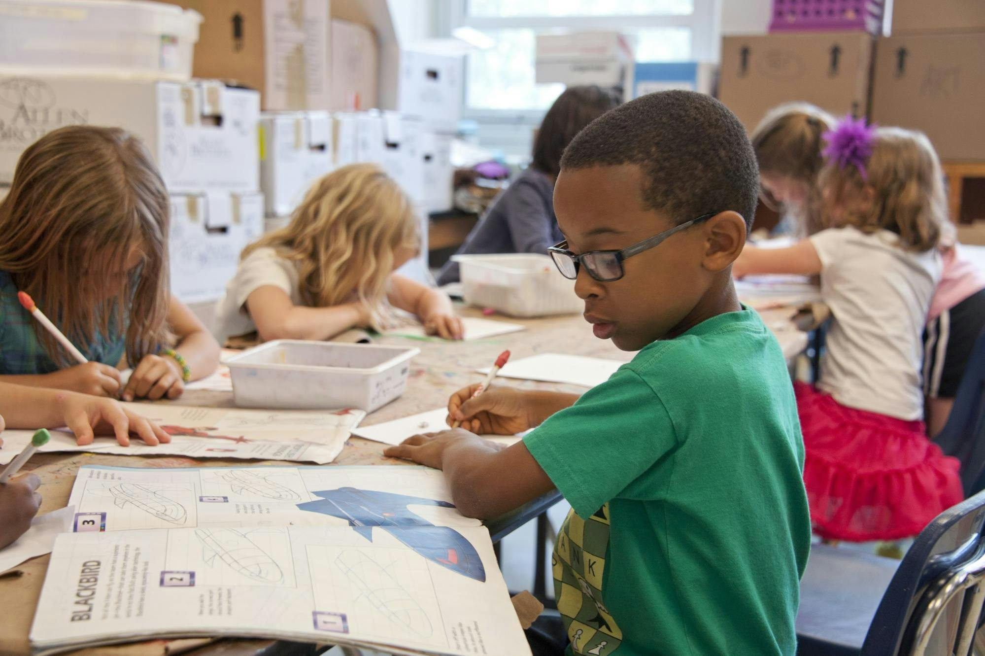 Young boy sits at a table with other students in the classroom, looking at a magazine and making notes.