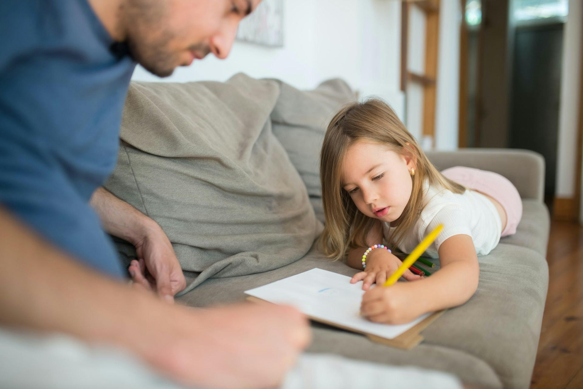 Young girls works with her father on writing activities on their couch.
