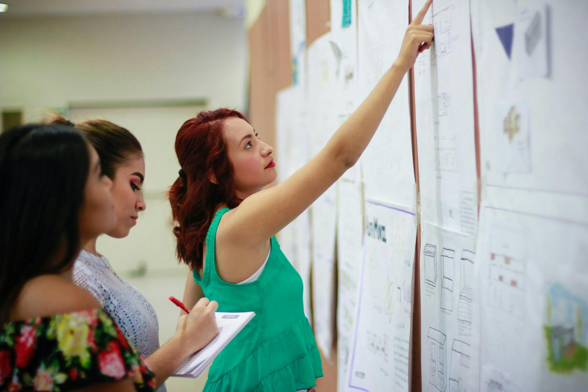 Female teacher points at charts on the wall while two other women take notes beside her.