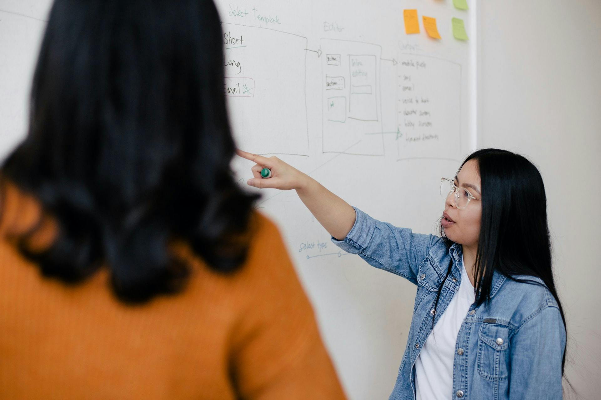 Two female students stand at a whiteboard and discuss a diagram.