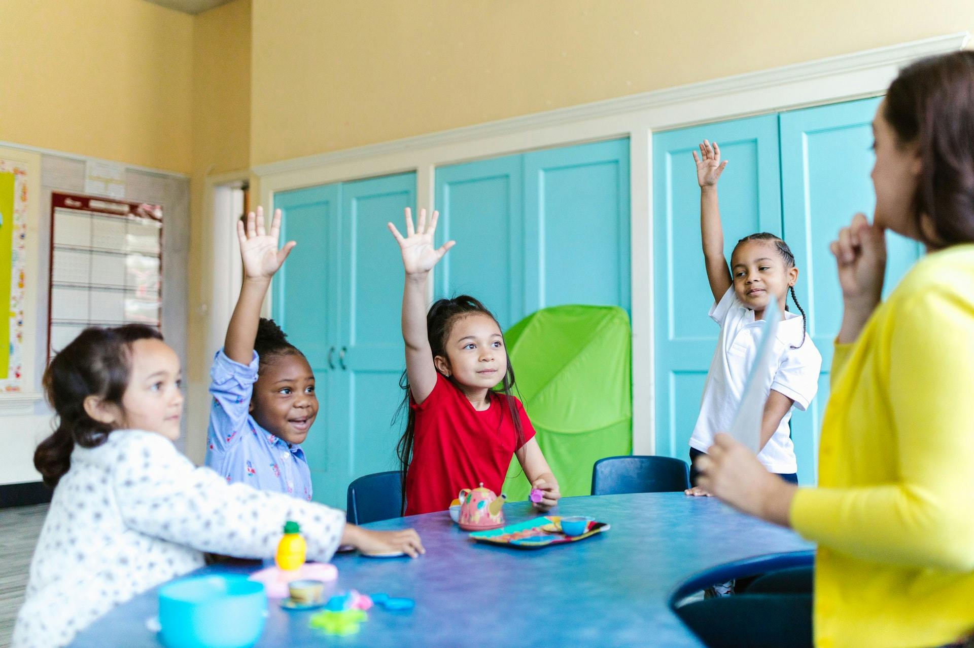 Group of students raise their hands around a table as they work on subtraction activities. 