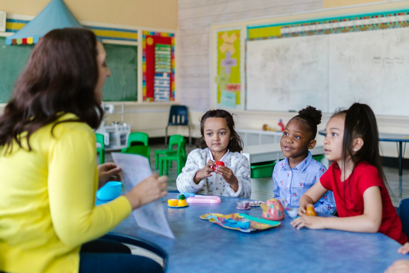 Three second-grade students sitting attentively at a classroom table, with their teacher across from them holding up a piece of paper for their lesson. 