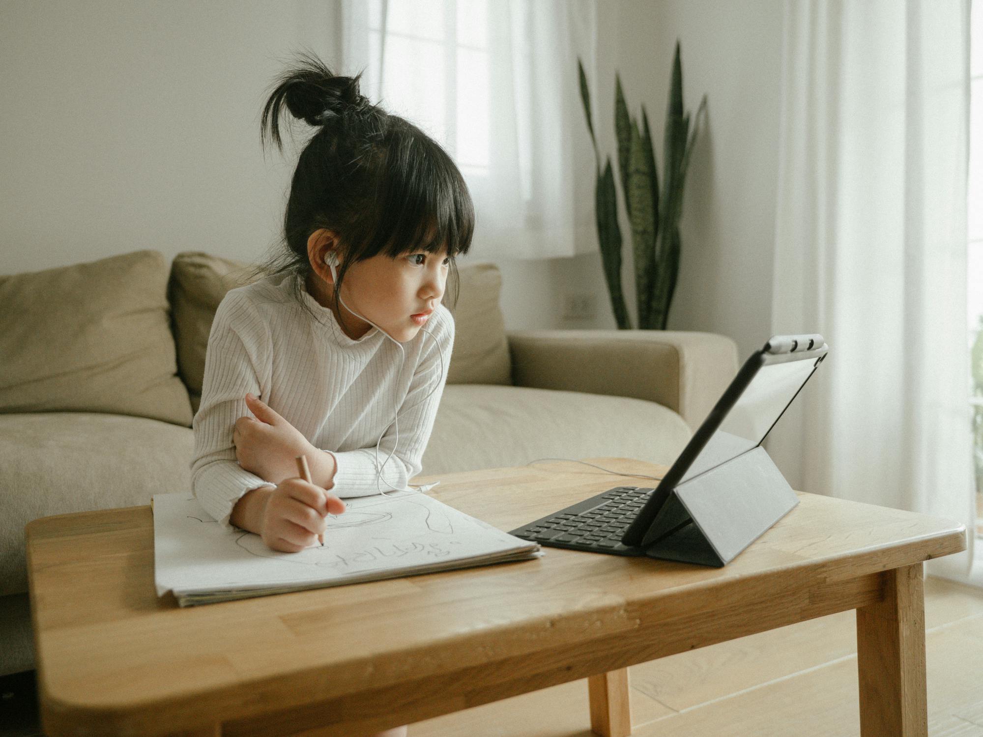 Young girl sitting at a table, watching something on her tablet while writing on a piece of paper with headphones in.