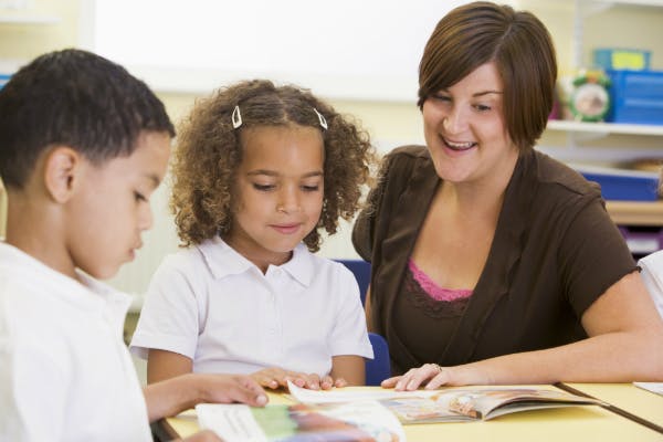 Teacher sits with two children as they read a book together.