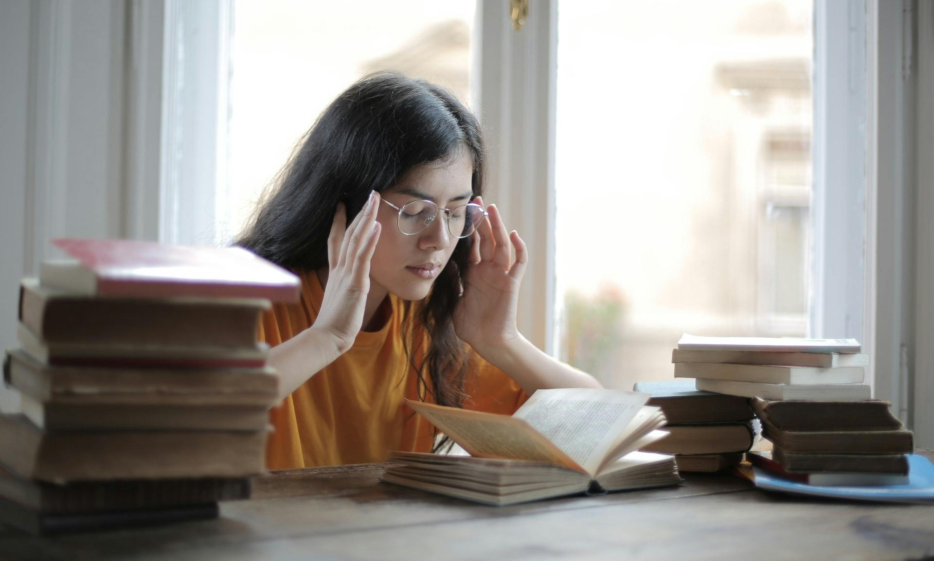 A high school student sits next to several piles of books and touches her temples as she struggles with rote memorization.