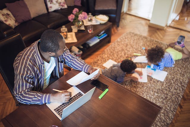 Father of two looks over at his kid doing homework on the carpet.
