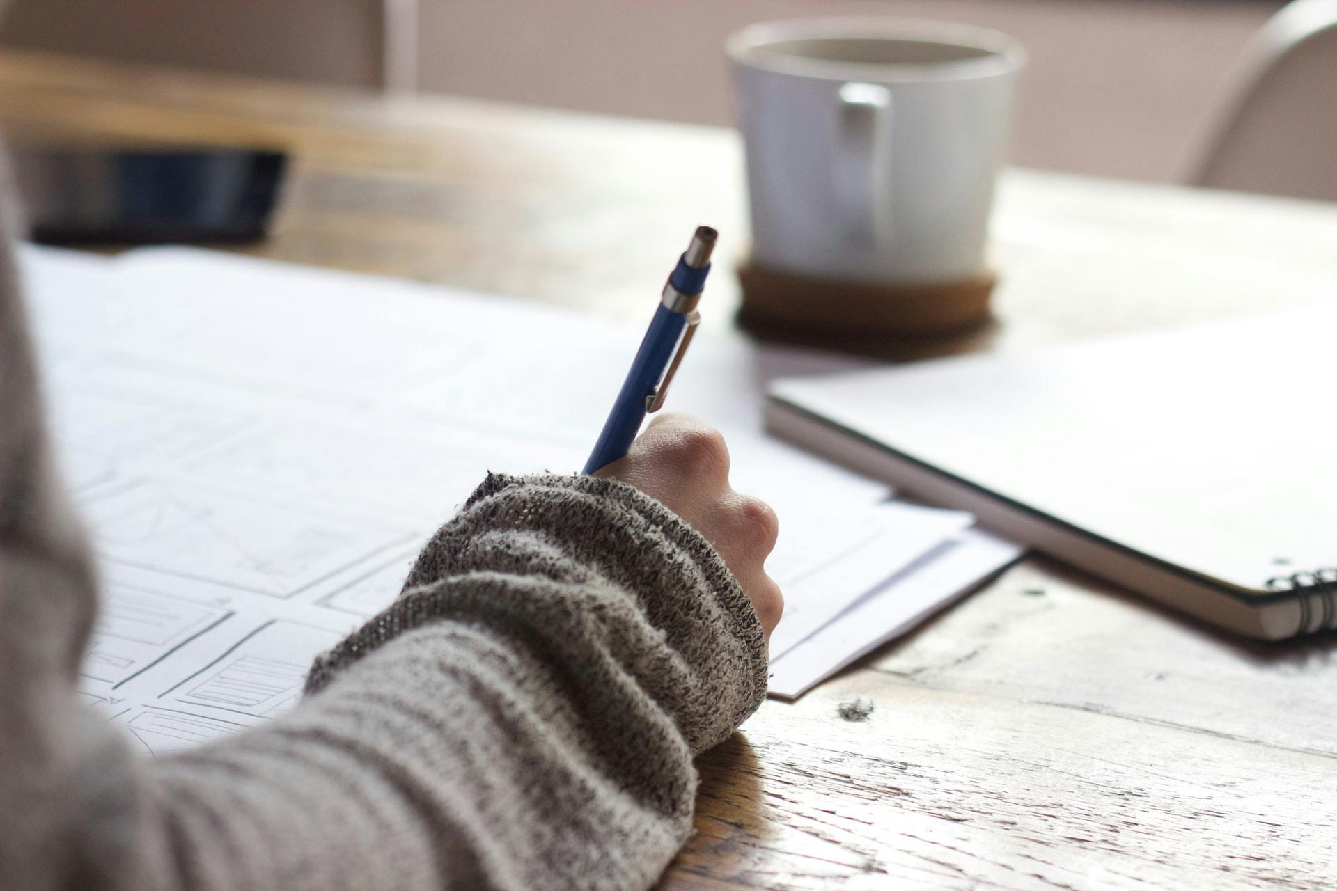 Teacher's hand writing a lesson plan with a notebook and a cup of coffee in the background.