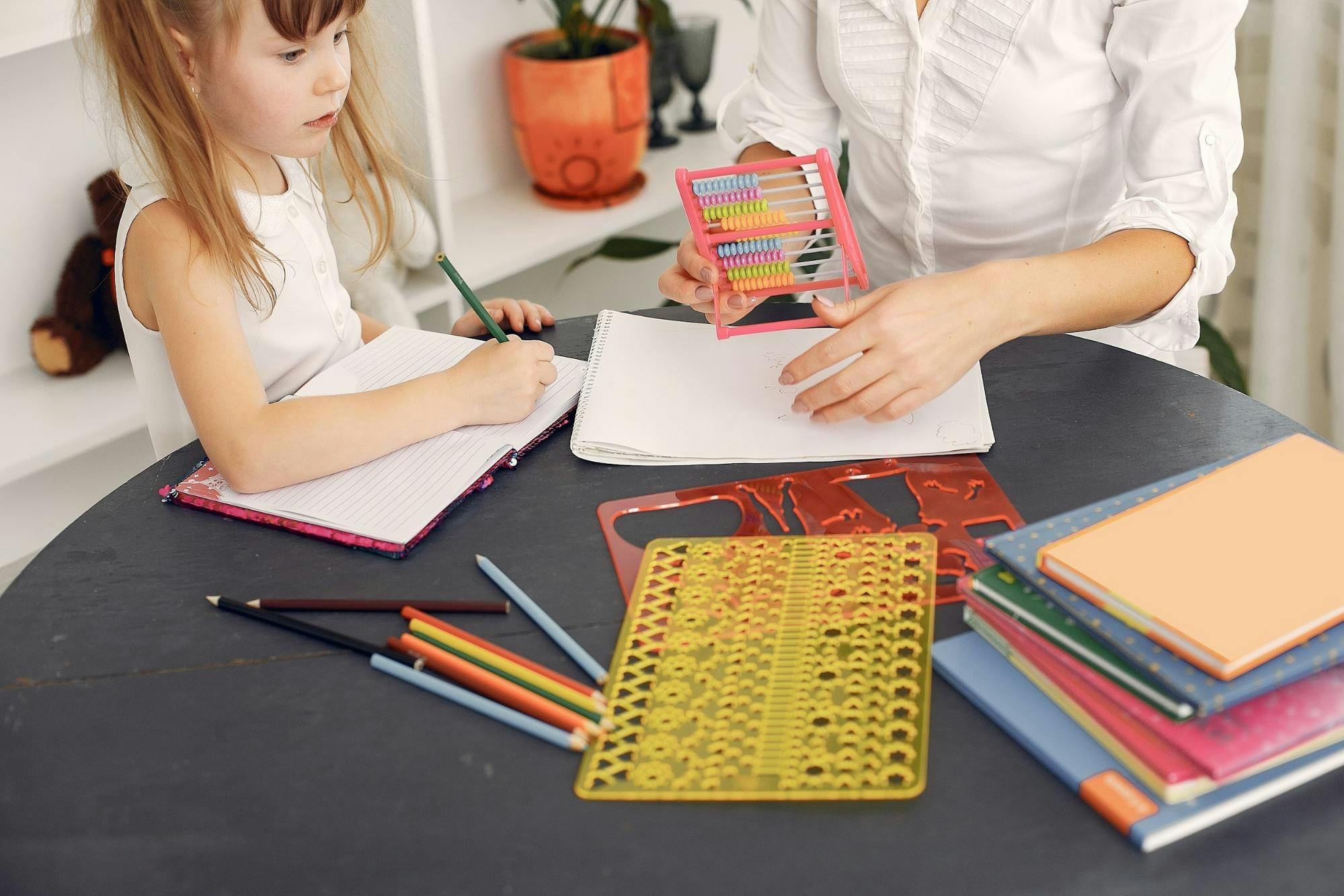 Young girls sits at a table with teachers, writing in a notebook and looking at writing supplies on the table.