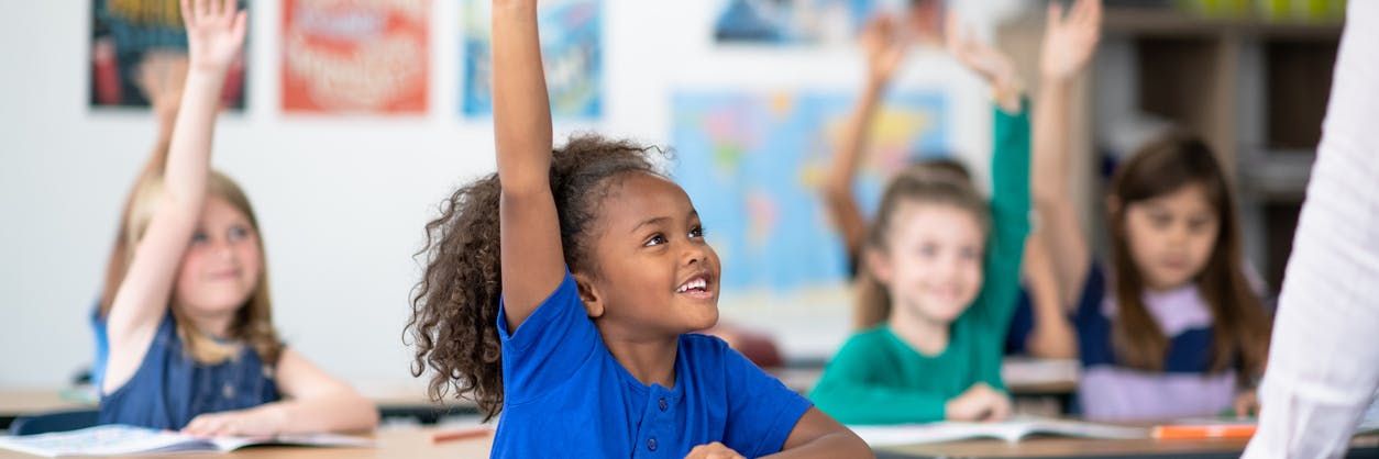 Second grade child smiling with her hand raised in class, with more students with their hands raised sitting behind her.