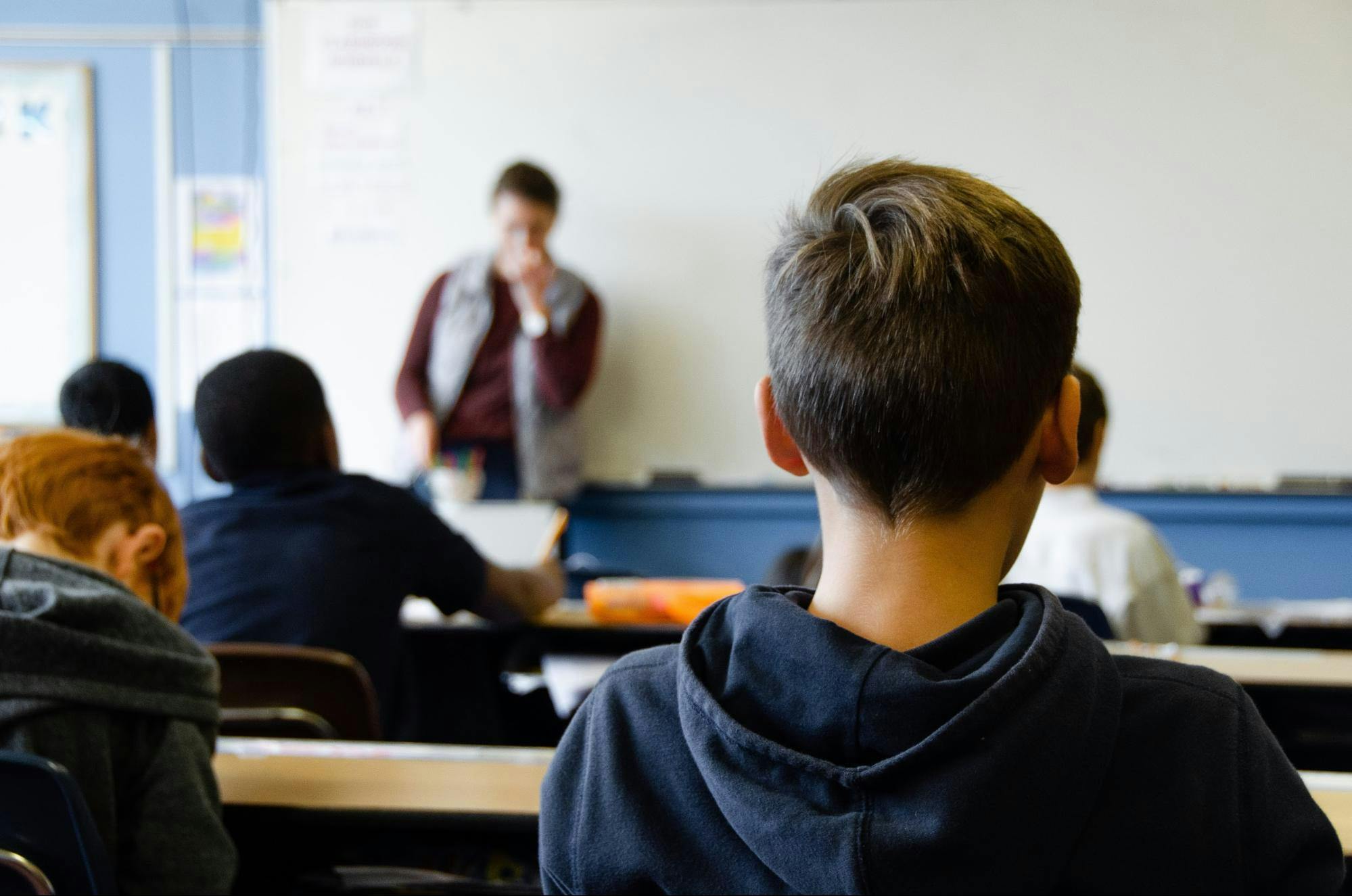 Students sit in a classroom working on social emotional activities and facing a teacher in front of a whiteboard.