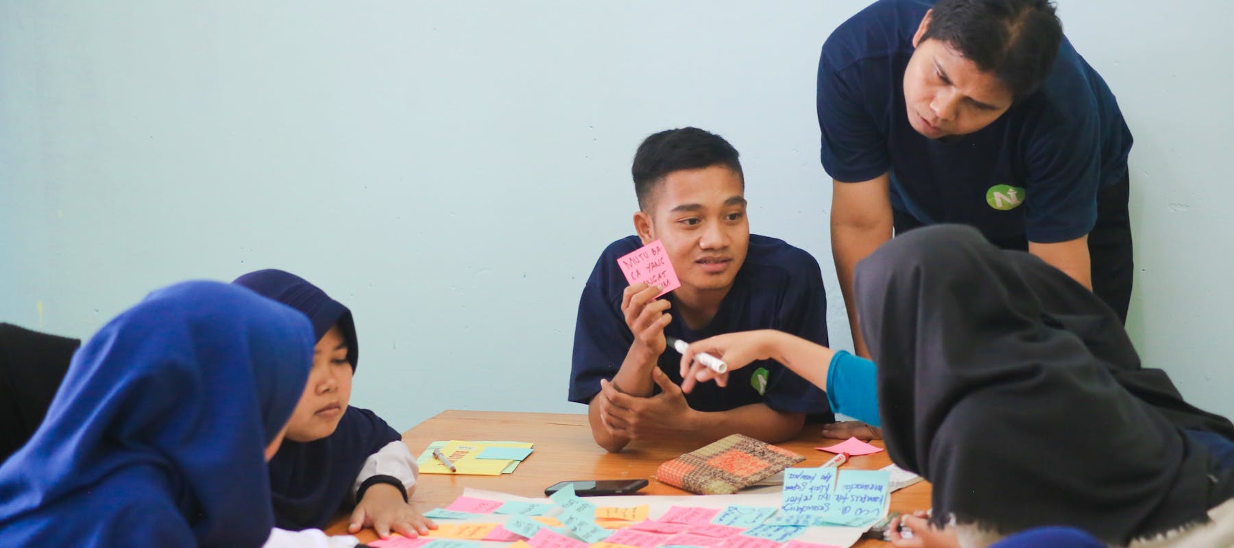 A classroom community gathers around a desk brainstorming ideas with post-it notes.