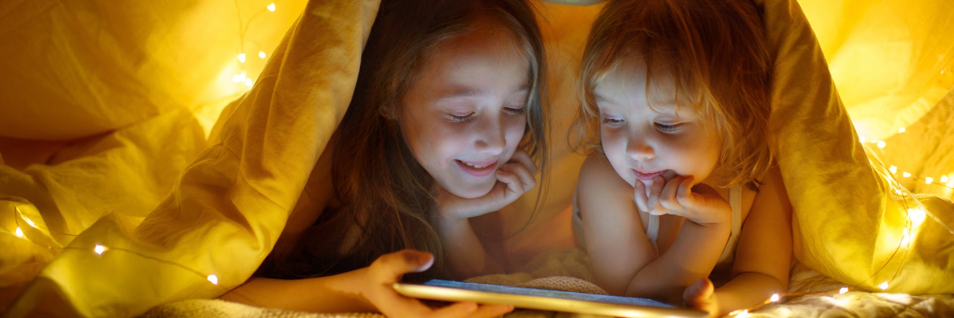 Two young children sitting in a blanket fort, exploring a reading website on their tablet device. 