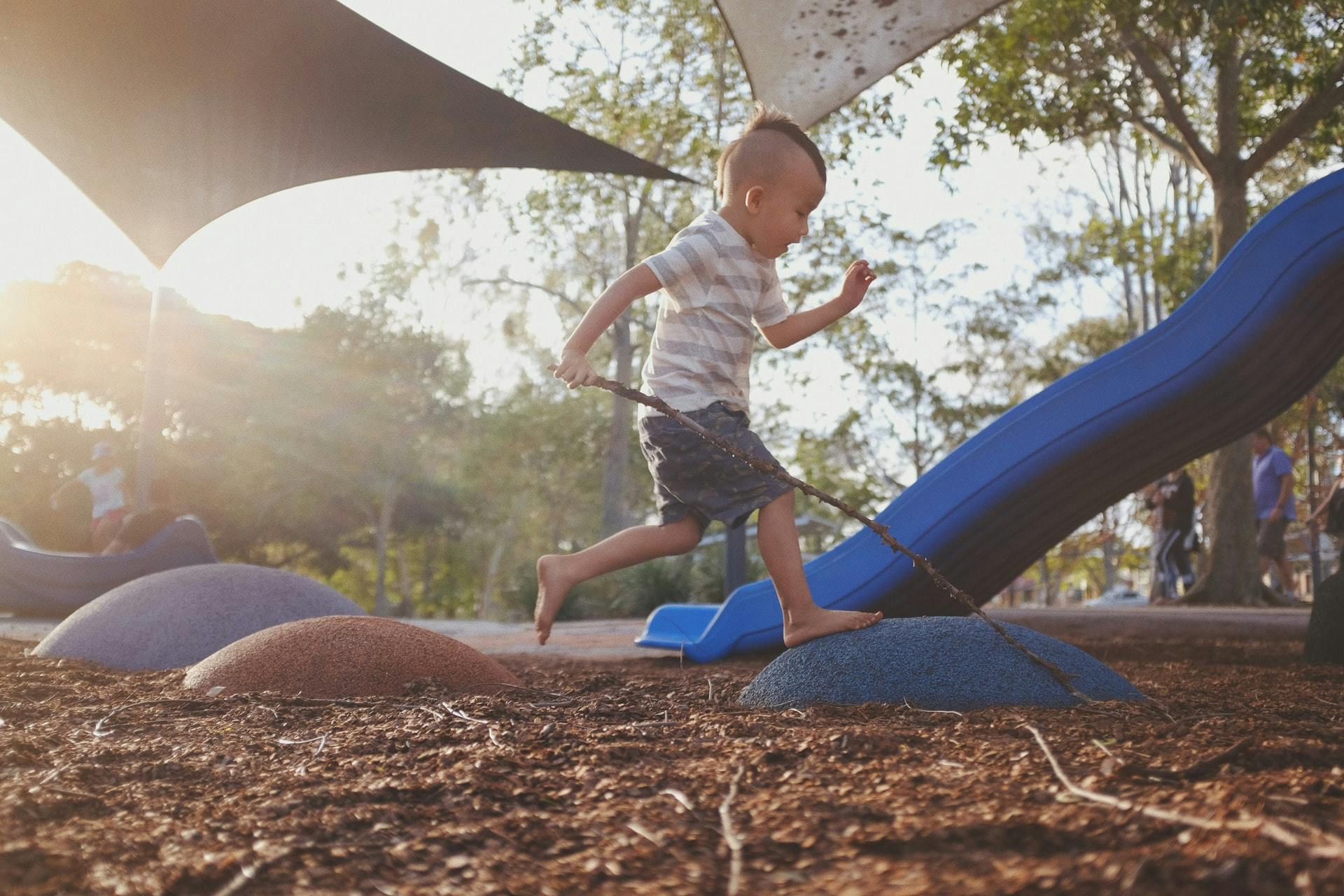 Young child plays outside on a playground while carrying a long stick.