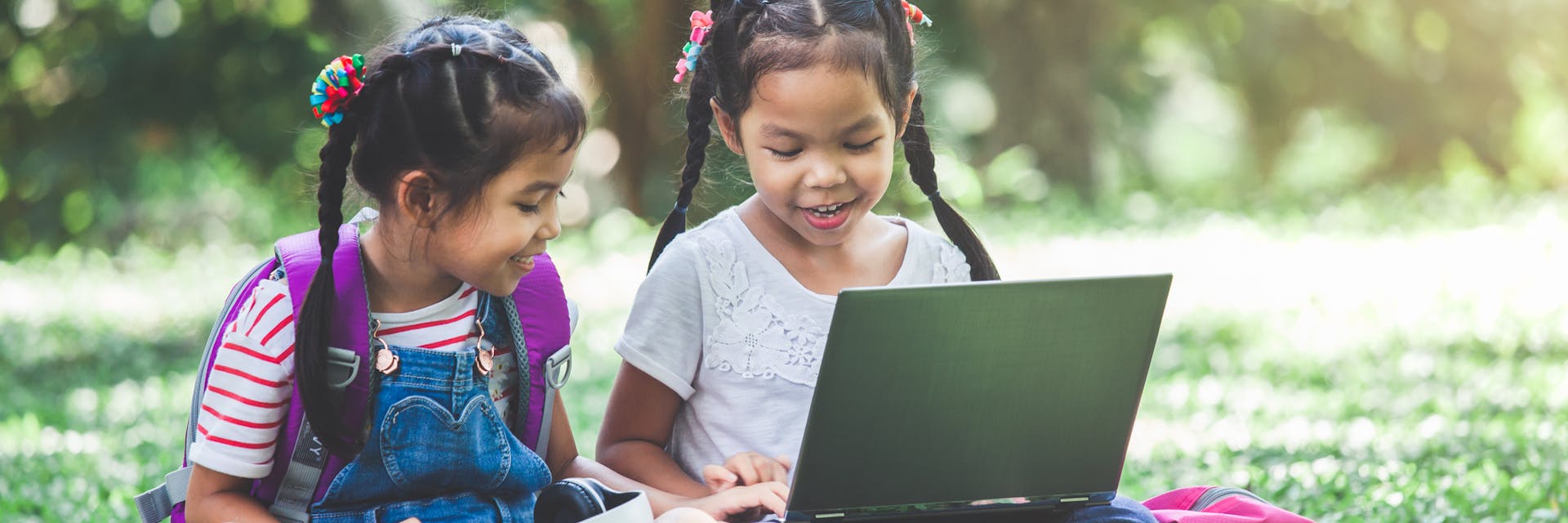 Two young girls sit outside and play Prodigy on a laptop to prevent summer learning loss.