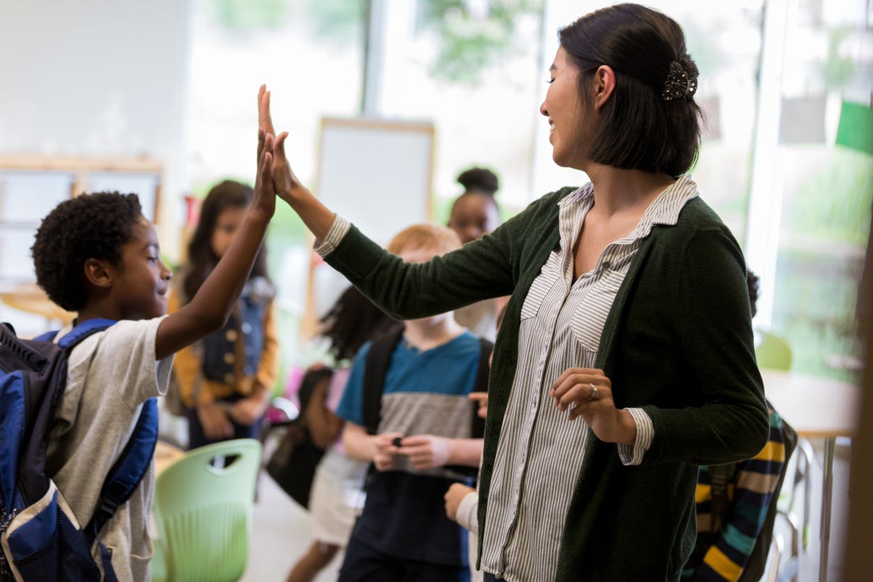 A teacher and their student high fiving and smiling with other students in the background.