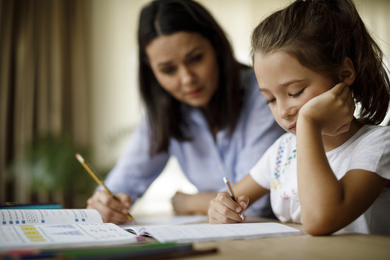 Bored young girl attempting to do her math homework with her mother sitting beside her.