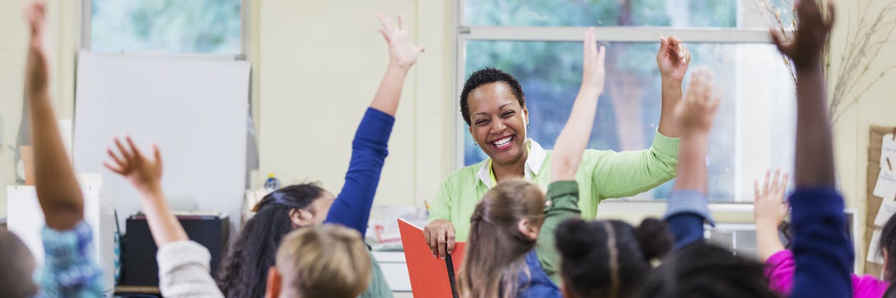A teacher smiles at her class as they eagerly raise their hands to answer a question of the day.