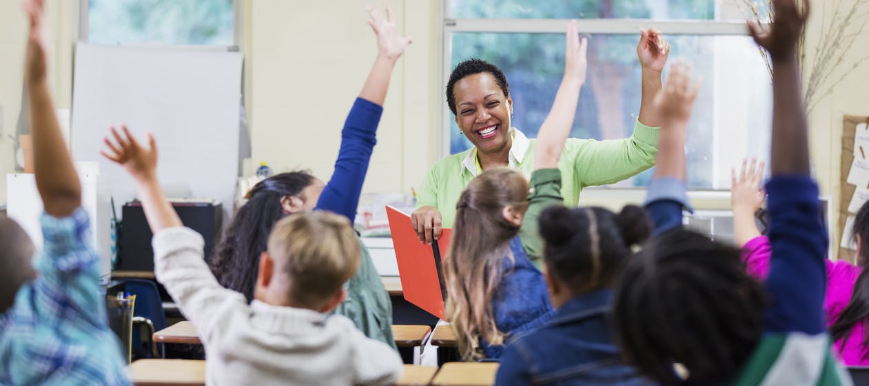 A teacher smiles at her class as they eagerly raise their hands to answer a question of the day.