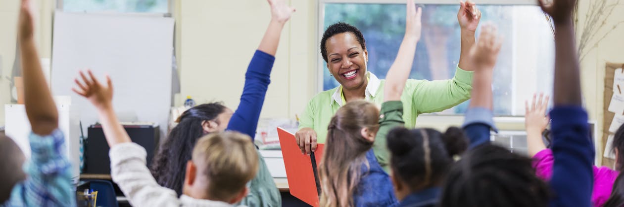 A teacher smiles at her class as they eagerly raise their hands to answer a question of the day.